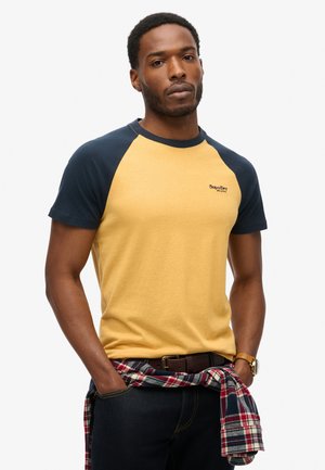 Yellow and navy short-sleeve T-shirt with raglan sleeves, featuring a small logo on the chest. Worn with a plaid shirt tied around the waist.