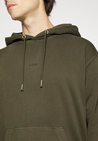 Olivgrön hoodie med dragsnören, med en subtil präglad logotyp. Slät tyg, känguruficka och ribbad mudd och fåll.
