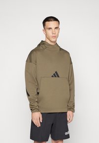 Felpa verde oliva realizzata in morbido tessuto, con tasca a marsupio e dettagli in logo neri. Abbinata a pantaloni corti neri per un contrasto.