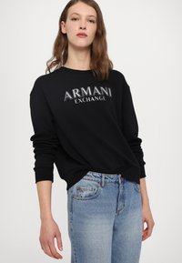 Zwarte sweatshirt met het logo "ARMANI EXCHANGE" in zilver, met ribgebreide boorden en zoom. Gecombineerd met lichtblauwe klassieke denim jeans.