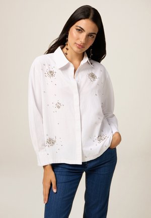 Witte overhemd met zilveren kraalversieringen aan de voorkant. Heeft een kraag en lange mouwen. Gepaard met donkerblauwe jeans.