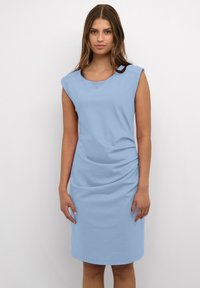 Frau mit langen braunen Haaren, die steht und ein ärmelloses hellblaues knielanges Kleid trägt, vor einem schlichten weißen Hintergrund.