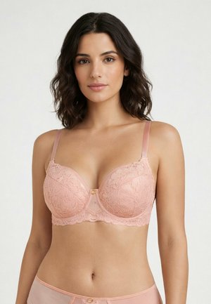 Kobieta w jasnoróżowym koronkowym biustonoszu i pasującym do niego bieliźnie, stoi na tle jednolitego jasnego tła, zwrócona twarzą do przodu.