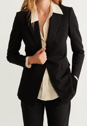 Femme portant un blazer noir cintré sur une chemise beige boutonnée, ajustant le blazer d'une main tandis que l'autre est dans la poche, vue du cou jusqu'au milieu des cuisses.