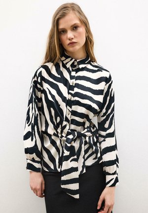 Jeune femme portant un chemisier boutonné à imprimé zèbre noir et blanc avec une taille nouée, debout devant un fond blanc uni.