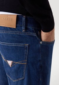 Jeans in denim blu scuro con cuciture gialle a contrasto. Presentano un'etichetta in pelle in vita e un logo triangolare sulla tasca posteriore.
