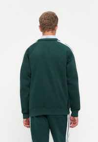 Green sweatshirt with a ribbed collar, white side stripes, and a relaxed fit. Features long sleeves and a smooth fabric texture.