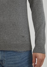 Maglione grigio realizzato in un tessuto morbido, con orlo e polsini a costine. Piccola etichetta logo sul lato inferiore. Indossato sopra jeans blu.