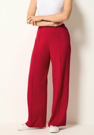 Donna con pantaloni rossi a vita alta e gamba larga con cuciture frontali abbinati a scarpe da ginnastica bianche e top bianco senza maniche.