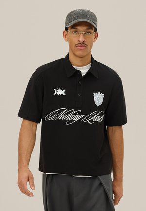 Młody mężczyzna w okularach i czapce nosi czarną koszulkę polo z napisem "Nothing Lasts" oraz ozdobnymi logo, w zestawieniu z ciemnymi spodniami na neutralnym tle.