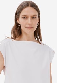 Top bianco a maniche corte con scollatura a barca, caratterizzato da bottoni dorati decorativi sulle spalle. Texture liscia, vestibilità comoda.