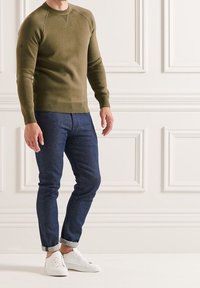Pull en coton vert olive avec poignets et ourlet côtelés, associé à un jean bleu foncé et des baskets blanches. Dispose d'un col rond et de manches raglan.