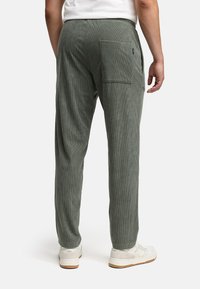 Mann in olivgrünen Cordhosen mit einer Gesäßtasche und weißen Sneakern, der vor einem schlichten weißen Hintergrund steht.