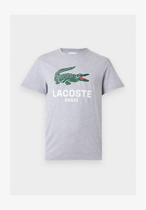 Svetlo sivo majico s kratkimi rokavi z zelenim logotipom krokodila in belim napisom "LACOSTE PARIS" na sredini prsnega koša.