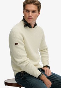 Cremfarbener gerippter Strickpullover mit Rundhalsausschnitt, getragen über einem dunkelkarierten Hemd, mit einem braunen Logopatch am Ärmel.