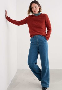 Femeie îmbrăcată într-un pulover de culoare ruginie, un gât de broderie cu dungi, blugi largi cu talie înaltă, stând cu o mână pe perete într-un decor neutru.