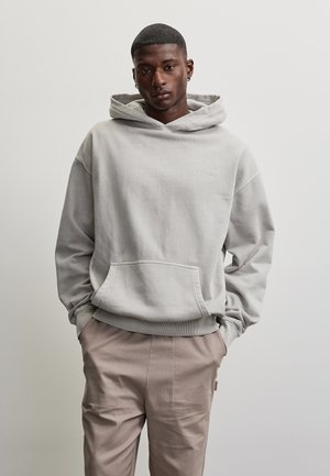 Jeune homme aux cheveux courts portant un sweat à capuche gris clair et un pantalon beige, debout les mains dans les poches devant un fond uni.