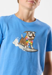 Ljusblå bomullströja med en tecknad hund som bär solglasögon och en regnbågsblomsterkrans, som surfar på en bräda med vågor.