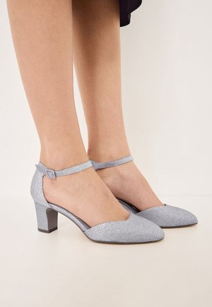 Des chaussures à talons hauts argentées pailletées avec une pointe fine, une bride de cheville et un large talon. Fini lisse et design découpé sur les côtés.