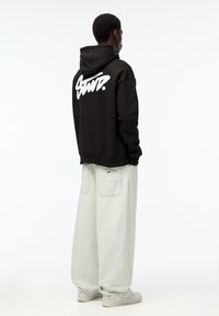 Černá mikina s bílým logem na zádech, v kombinaci se světle šedými širokými kalhotami. Modelka stojí s otočeným tělem, má na sobě šedé tenisky.