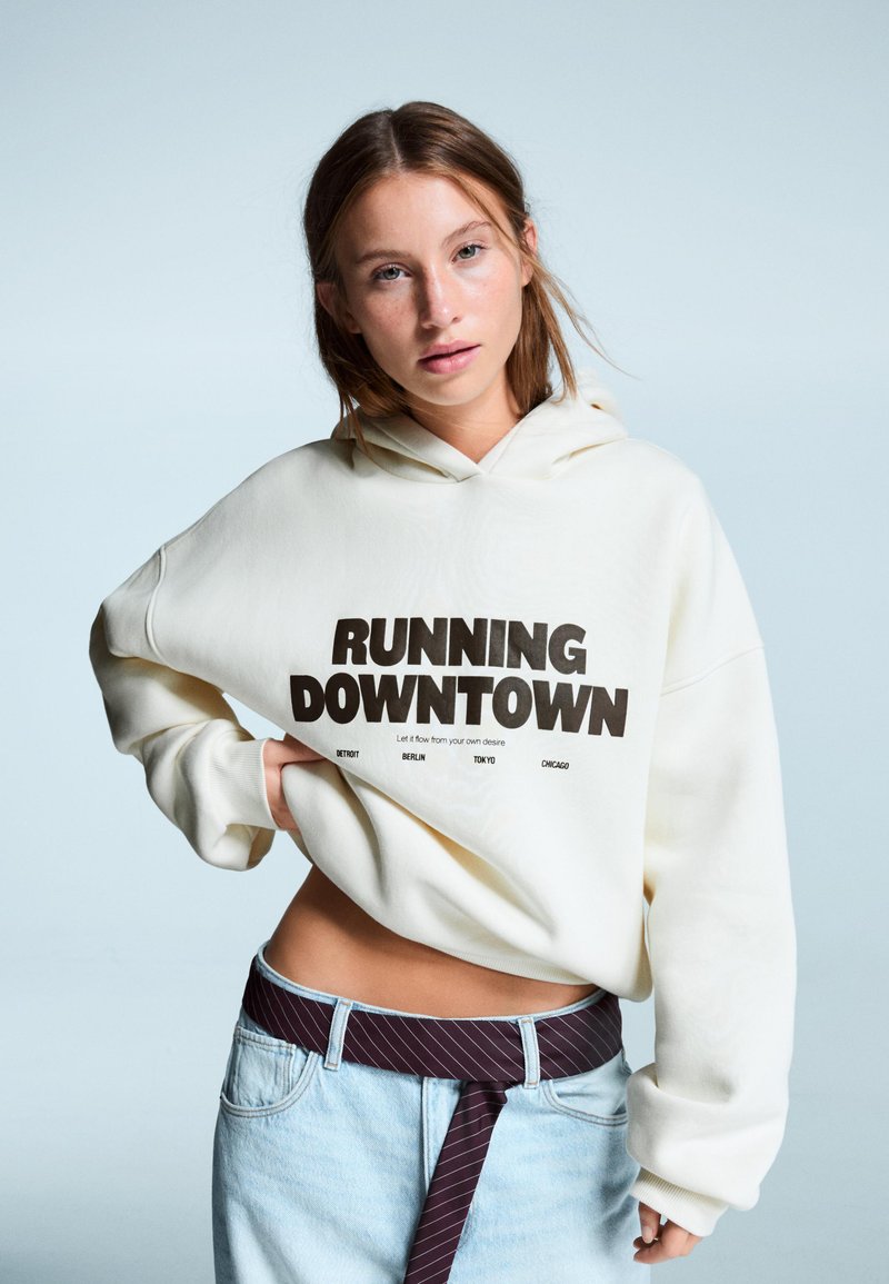 Krémová oversized mikina s nápisom "RUNNING DOWNTOWN" v tučnom čiernom písmene, skombinovaná s svetlými denimovými džínsami a vzorovaným opaskom.