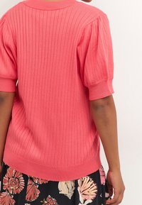 Korálový úpletlý svetr s krátkými nabíranými rukávy, zakončený širokým žebrovým lemem. Nosený přes sukni s květinovým vzorem.