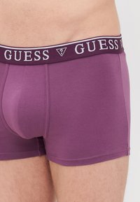 Violet bokseršorti, kas izgatavoti no mīkstas auduma ar pieguļošu dizainu. Elastīgā josta ar zīmola nosaukumu "GUESS" baltā krāsā tumšā joslā.
