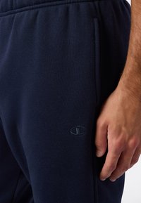 Męska ręka spoczywająca w pobliżu kieszeni granatowych spodni dresowych z małym, haftowanym logotypem Champion na udzie.