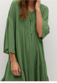 Abito verde a strati realizzato in tessuto strutturato con una vestibilità ampia, caratterizzato da una scollatura annodata e maniche larghe per una silhouette rilassata.
