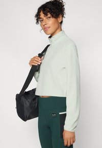 Hellgrüner, kurz geschnittener Fleece-Pullover mit hohem Kragen, kombiniert mit dunkelgrünen Leggings mit einer Seitentasche und schwarzen Akzenten. Schwarzer Schulterriemen.