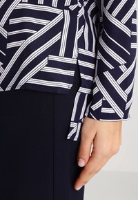 Mörkblå topp med vitt randigt mönster, med en något avkortad design, långa ärmar och sprund på sidorna för bättre rörelsefrihet.