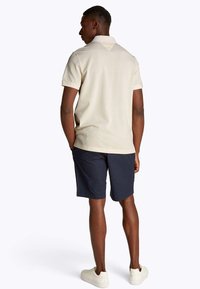 Polo beige con colletto testurizzato, abbinata a pantaloncini blu navy. Scarpe da ginnastica bianche completano l'outfit, evidenziando un look casual.