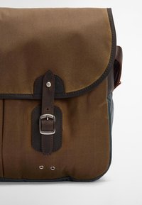 Καφέ καμβάς τσάντα μηνυμάτων με καπάκι, δερμάτινες λεπτομέρειες και μεταλλικό κούμπωμα. Διαθέτει δύο ορατές θηλιές μπροστά.