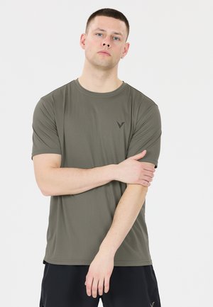 Mand med kort hår iført en olivengrøn t-shirt og sorte shorts, stående med den ene arm krydset over kroppen.