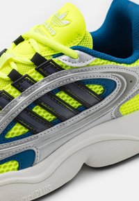 Heldere gele Adidas sneaker met zwarte strepen, zilveren details, blauwe voering en dikke witte zool met "adiPLUS" merknaam.