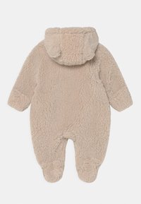 Combinaison en polaire beige avec un extérieur texturé et duveteux. Dispose d'une capuche avec des oreilles arrondies et de doux poignets aux poignets et aux chevilles pour plus de chaleur.