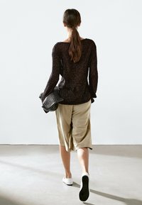 Femme aux longs cheveux attachés en queue de cheval, marchant de dos, portant un pull en tricot foncé, un short beige à jambes larges, des chaussures blanches et tenant un sac noir texturé.