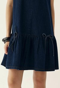 Denim kleita ar izplūdušu apakšmalu, kas dekorēta ar krošņu detaļām un regulējamiem saitēm sānos. Tumši zila krāsa ar kontrastējošām šuvēm.