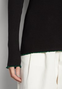 Top nero a maniche lunghe con righe e dettaglio ondulato verde all'orlo, abbinato a pantaloni bianchi plissettati, che presentano un design aderente e testurizzato.