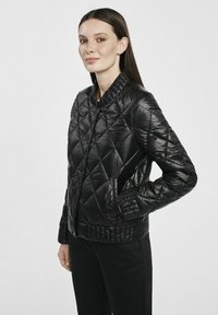 Czarna pikowana kurtka z prążkowanym kołnierzem i mankietami, z bocznymi kieszeniami i gładką, błyszcz ąca teksturą. Noszona z czarnymi spodniami.