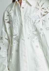 Chaqueta blanca de camisa con intrincados bordados florales y acentos de perlas, cierre frontal con botones, cuello y cordón en el dobladillo.