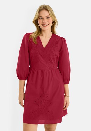 Mujer sonriente que lleva un vestido rojo hasta la rodilla, de manga larga, con escote en V y bordado con ojales, de pie frente a un fondo liso y claro.