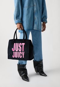 Fekete velúrból készült tote táska rózsaszín "JUST JUICY" hímzéssel, kerek fogantyúkkal és strukturált formával, farmer ruházattal és fekete csizmával párosítva.