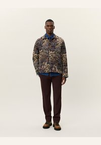 Floral patterned jacket with shades of purple, beige, and green; paired with a blue shirt and maroon trousers; brown shoes with laces.
