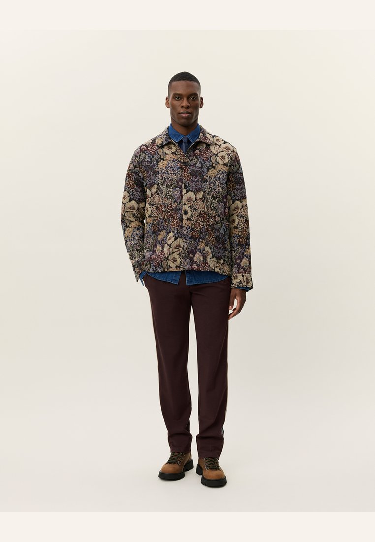 Floral patterned jacket with shades of purple, beige, and green; paired with a blue shirt and maroon trousers; brown shoes with laces.