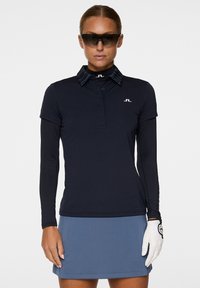 Granatowa koszulka polo z długim rękawem, z kołnierzykiem, w paski w okolicy kołnierza oraz logo na piersi; zestawiona z jasnoniebieską spódnicą i białą rękawicą golfową.