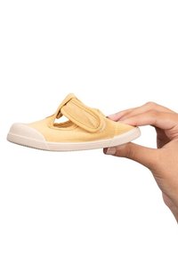 Zapato de ante amarillo con tira de velcro, punta redondeada, suela de goma crema y superficie texturizada; sostenido en una mano frente a un fondo blanco.
