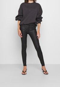 Svarta läder skinny jeans i kombination med en mörk oversized sweatshirt. Modellen bär svarta flip-flop sandaler. Slät textur, figurnära design.