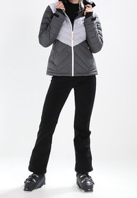 Vadderad skidjacka i grått och vitt med ett chevronmönster och dragkedja. Kombination med svarta byxor och skidstövlar.