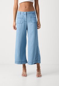 Žena oblečená v světlých modrých širokých kalhotách do výšky kotníků se předními náprsními kapsami a béžovými sandály na podpatku na bílém pozadí.